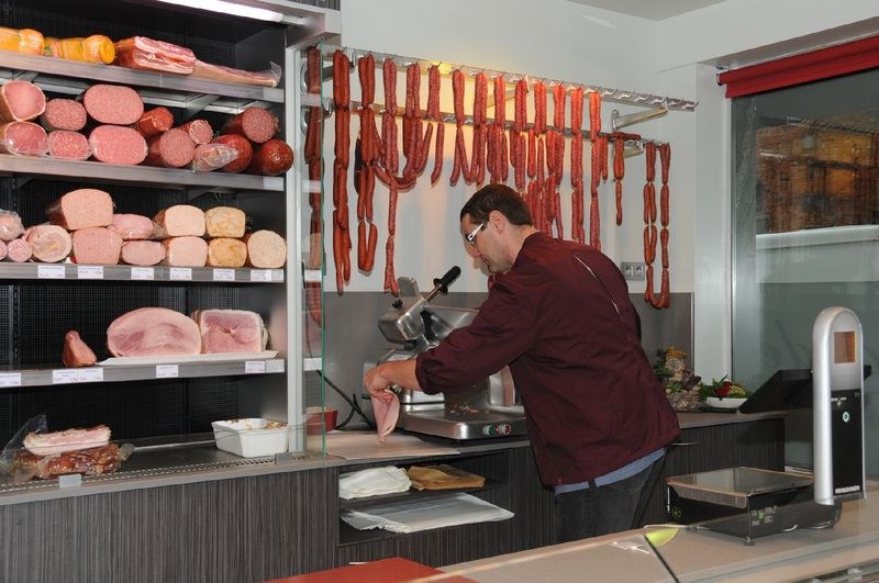 Stéphane tranche du jambon derrière le comptoir, devant les charcuteries suspendues